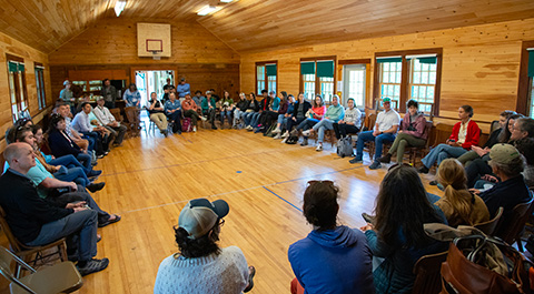 A group of people sitting in chairs around an indoor wooden basketball court.