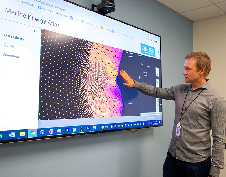 A man gesturing at a large screen that shows a visualization of wave energy resources off the Pacific coast
