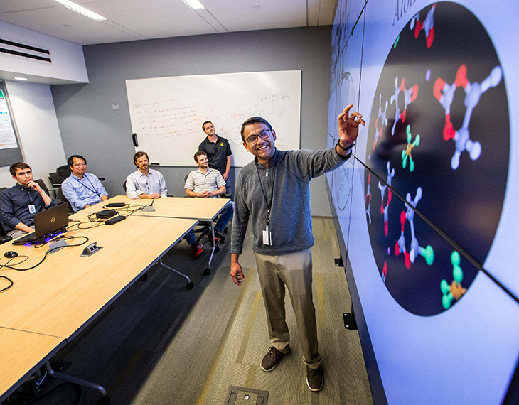 A researcher stands in front of a large screen. Five other researchers sit in the background.