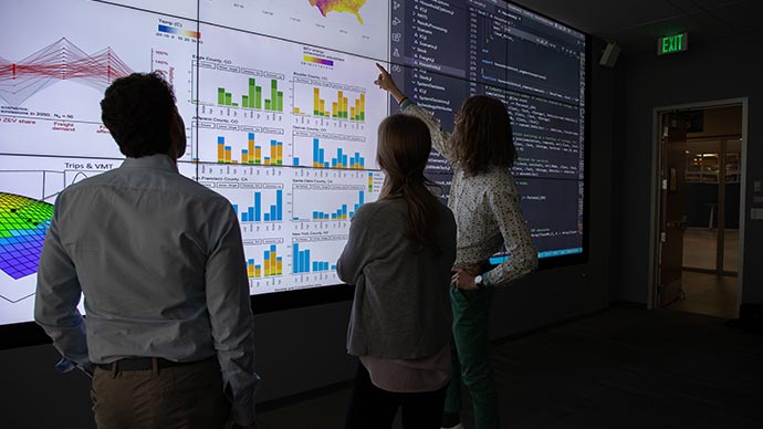 Researchers view charts and graphs on an illuminated wall display, analyzing transportation and mobility scenarios.
