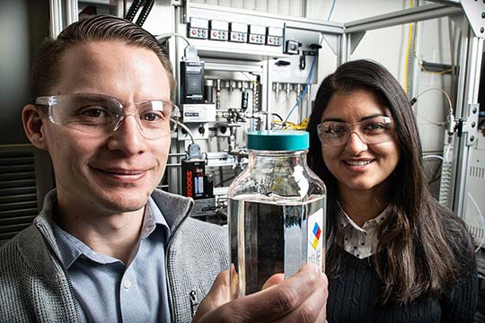Photo of two people in laboratory setting. One person is holding a container of clear liquid.