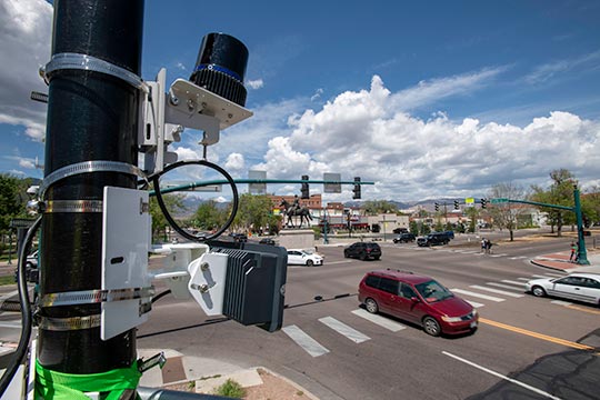 Cars and pedestrians at a four-way intersection with stoplights. Scientific equipment is strapped to one of the stoplight posts.