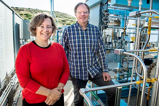 A woman and man smile at the camera as they stand by scientific equipment.