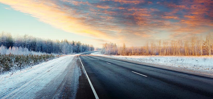 A rural road surrounded by snow and trees during sunset.