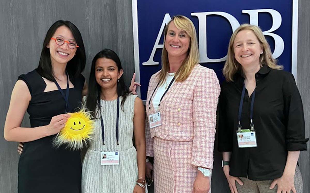 Four women smiling for a photo in front of a large sign. The woman on the far left is holding a small, fuzzy, yellow sun stuffy.