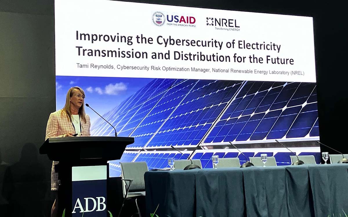 A woman presenting at a podium with an empty table behind her and a large branded slide in the background.