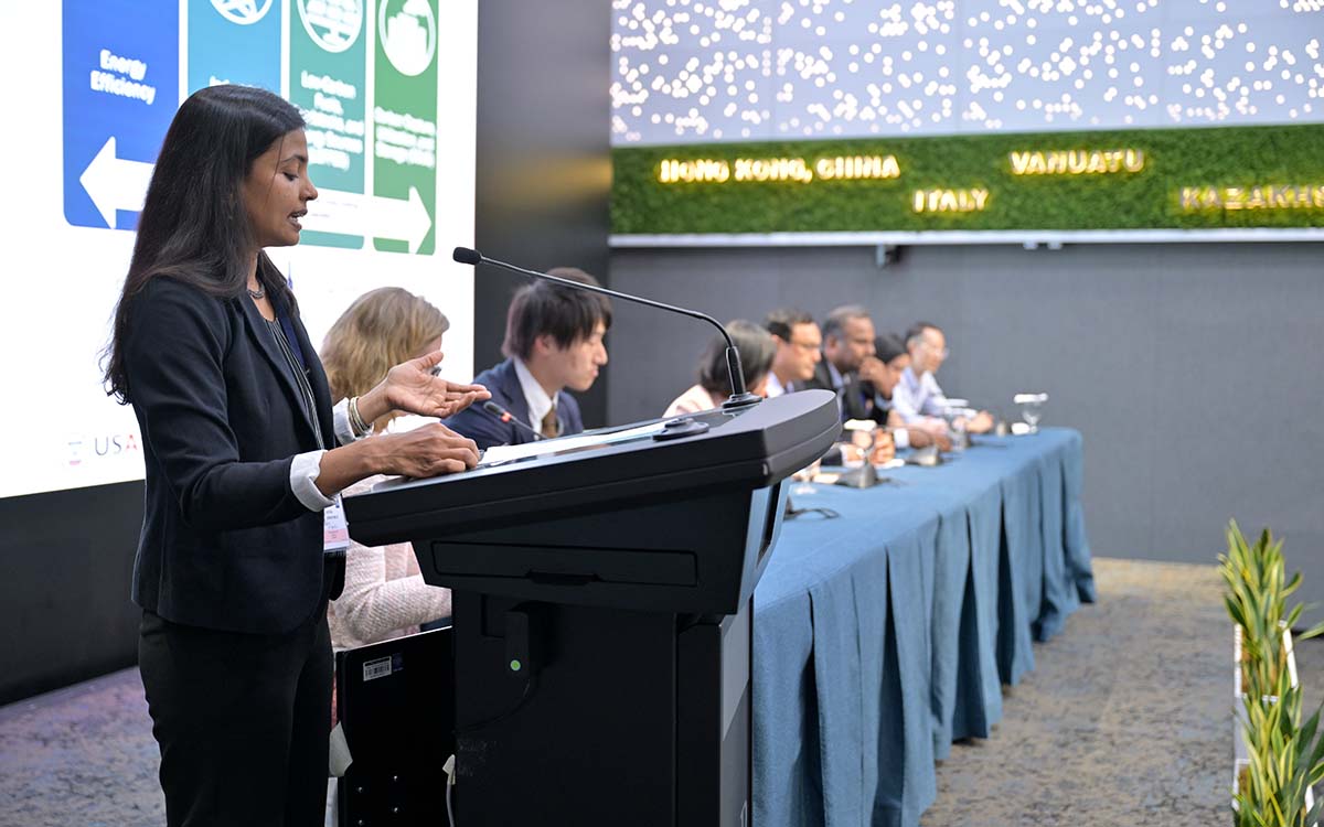 A side angle view of a woman presenting at a podium, with several individuals seated at a long table behind her.