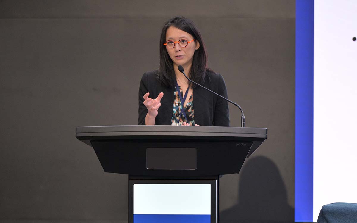 A woman in glasses presenting at a podium with one of her hands raised. People are seating in the foreground.