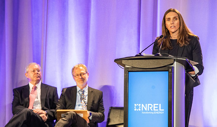 A woman speaking at a podium. Two men sit behind her.