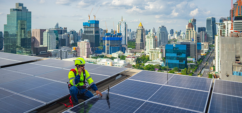 A worker in a construction outfit and hard hat uses a laptop and phone on a rooftop covered with solar panels. In the distance are skyscrapers, trees, a road, and clouds.