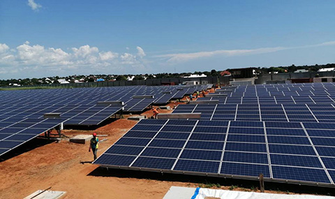 A field of solar panels stretches to the horizon, with trees and buildings in the distance. A worker with a construction vest and hard hat walks between the panels.