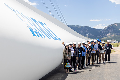 A group of individuals in hard hats smile and give a thumbs up as they stand in front of a large wind turbine blade with the NREL logo on it.