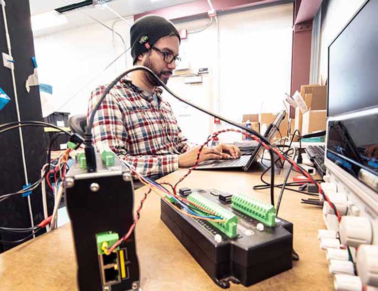 Aidan Bharath sits at a laptop next to wires and electronics.