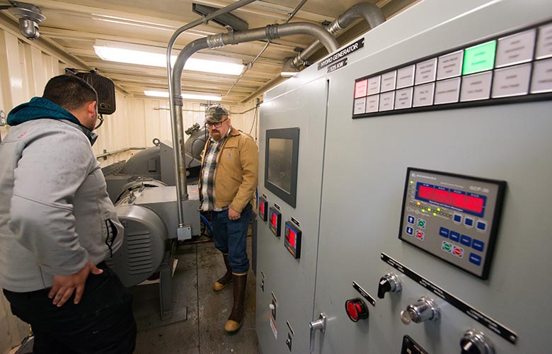Two men in winter coats stand next to a large electrical box in a facility.