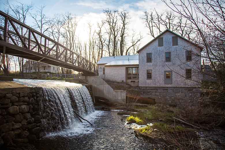  A small dam creates a waterfall under a foot bridge next to an old house.