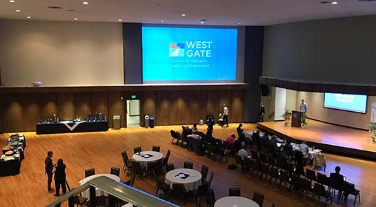 Image of an auditorium that has round tables, an audience sitting in chairs, and a person at a podium on a stage