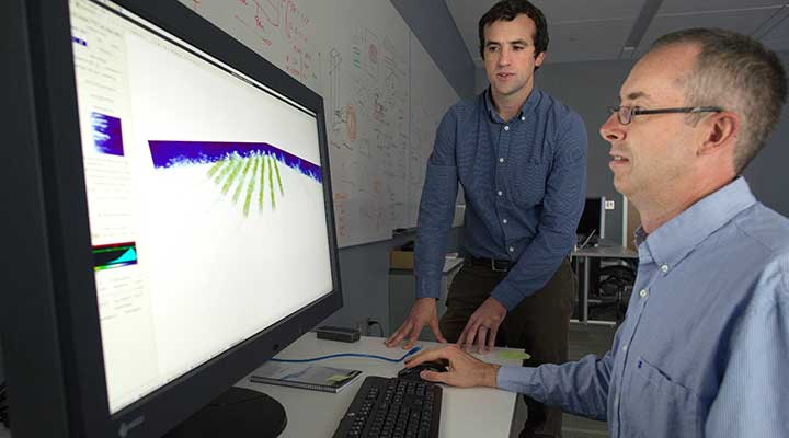 A photo of two men looking at a computer monitor that displays blue and green wind-modeling graphics.